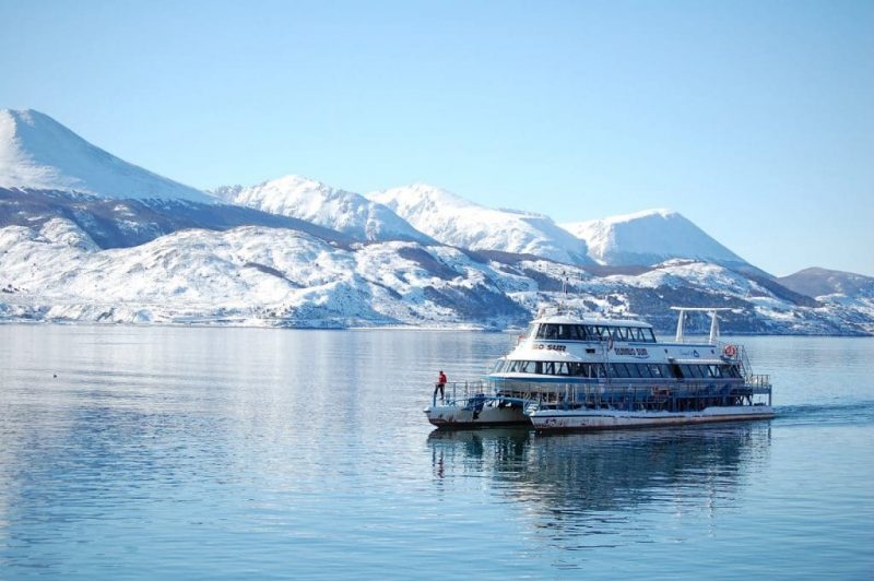 Barco navegando pelo Canal Beagle em Ushuaia, nota-se montanhas cobertas de neve ao fundo da imagem