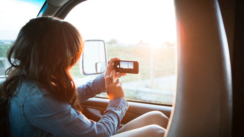 Mulher filma a paisagem pela janela de um carro em seu celular