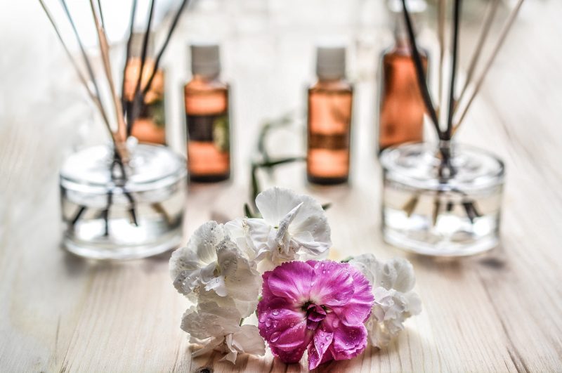 Imagem ilustrativa de perfumes, frascos de perfumes ao fundo e flores à frente