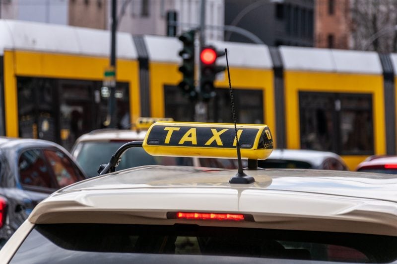 Parte superior de um táxi, dando destaque para a placa com o nome TÁXI, em uma rua movimentada aparentemente. Nota-se um semáforo ao fundo com a luz vermelha acesa.
