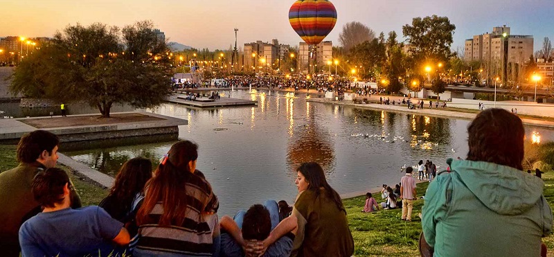 Turistas em um parque em Mendoza, nota-se um balão ao fundo e um lago no centro da imagem com pessoas ao redor conversando e passeando