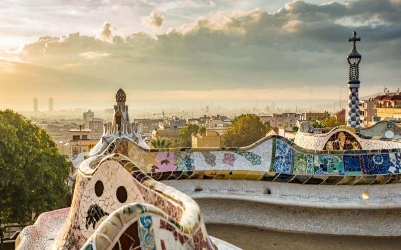 Vista do Park Güell de frente para a cidade de Barcelona. O céu está com nuvens e a luz do sol é bem leve.