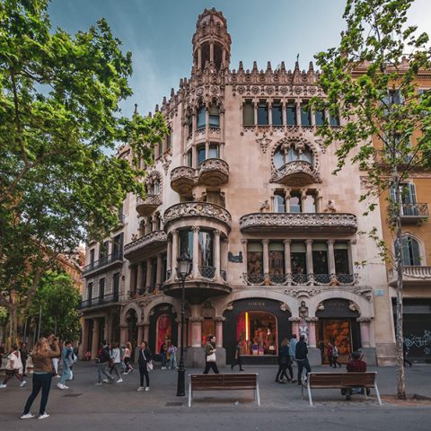 Vista de prédio em Passeig de Gràcia em Barcelona. Percebe-se pessoas transitando em frente à loja, duas árvores (cada uma ao lado) da loja e bancos do lado direito de um poste em frente ao prédio.