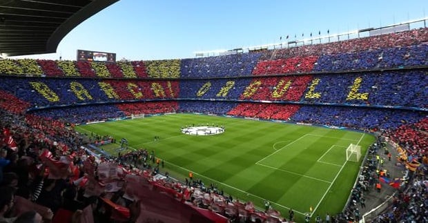 Estádio do Barcelona em jogo. O campo está vazio, mas as arquibancadas estão cheias e coloridas com as cores do time: azul, amarelo e vermelho. Ainda, nas arquibancadas formam-se os dizeres "Barcal Orgull" em amarelo.