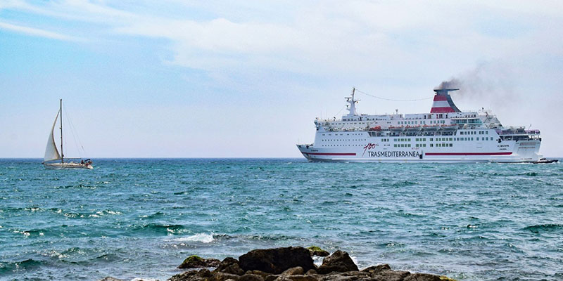 Ferry Boat Transmediterranea no mar. Há um barco a vela do lado esquerdo do ferry e há algumas rochas em primeiro plano na imagem