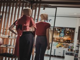 Dois manequins com roupas femininas de frente para a vitrine de uma loja. Os manequins vestem uma blusa vermelho escuro e calças escuras. Um deles está com a mão esquerda apoiada na cintura.