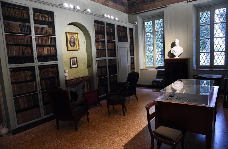 Interior do Museu Alessandro Manzoni. O cômodo tem prateleiras de livros, cadeiras e mesas, além de um busto de um homem ao lado de uma janela.
