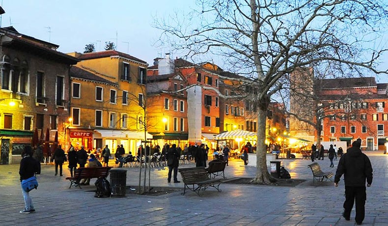 Praça na Região de Dorsoduro em Veneza. Nota-se os estabelecimento com as luzes acesas, pois está anoitecendo, e pessoas caminham pela praça com alguns bancos. Do lado direito da imagem há uma árvore sem folhas.