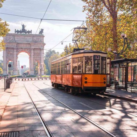 Vista do Arco della Pace em Milão. Ele está ao fundo e há um bondinho elétrico à frente, mais próximo ao primeiro plano da imagem. A cidade está pouco movimentada e está ensolarado.