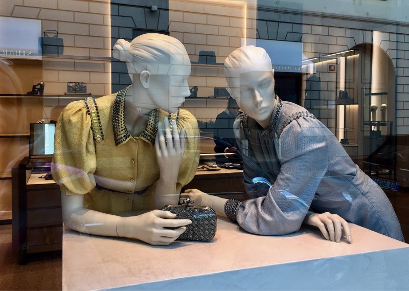 Dois manequins posicionados em loja na Via della Spiga. Os manequins femininos estão posicionados de modo que simulam uma conversa. O manequim à esquerda veste uma camisa com mangas bufantes na cor amarela e o manequim à direita veste uma camiseta na cor cinza com mangas que chegam aos pulsos.