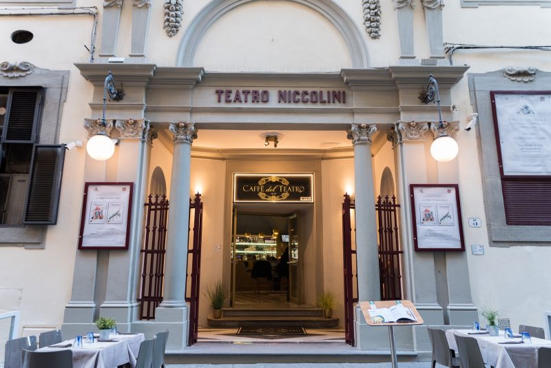 Fachada do Teatro Niccolini em Florença. A estrutura antiga do lugar tem muitos detalhes (alguns na cor vermelha) é pintada em cores claras e há quadros com informes na entrada.