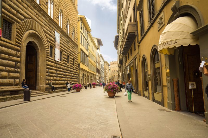 Vista da Via Tornabuoni em Florença. Nota-se prédios com o estilo vintage e vasos com flores rosa espalhados pela rua.