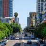 Road with multiple parked cars, green trees, modern buildings along it, sunny day in San Diego, USA